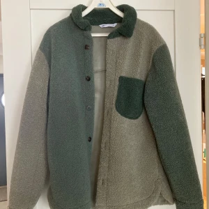 Grön teddyjacka från Zara - Säljer en mysig grön teddyjacka från Zara Man med knappar framtill och en praktisk bröstficka. Jackan har en skön krage och långa ärmar. Den är i en härlig tvåfärgad design med olika nyanser av grönt. Använd ca 3 ggr