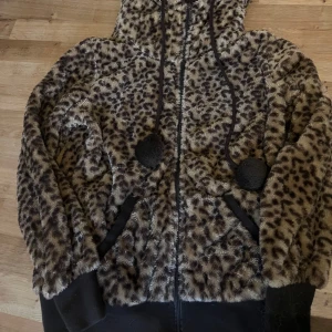 Leopardmönstrad teddyjacka från Benter - Mysig leopardmönstrad teddyjacka från Benter med huva och dragkedja. Jackan har långa ärmar och dekorativa öron på huvan. Perfekt för att hålla sig varm och stilren under kyliga dagar.