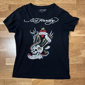 Ed Hardy T-shirt Black - Snygg svart t-shirt från Ed Hardy med ett coolt tryck av en dödskalle och texten 'New York City'. Perfekt för dig som gillar en edgy stil. T-shirten har korta ärmar och är gjord i bomull för en skön passform. Storlek mer som M