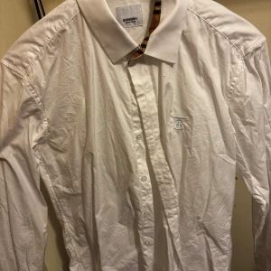 Vit skjorta från Burberry - Säljer en stilren vit skjorta från Burberry med klassiskt rutigt mönster på insidan av kragen och ärmsluten. Skjortan har långa ärmar och en broderad logotyp på bröstet. Perfekt för en elegant look.