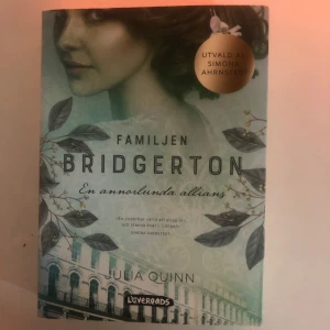 Familjen Bridgerton: En annorlunda allians - Boken 'Familjen Bridgerton: En annorlunda allians' av Julia Quinn, utvald av Simona Ahrnstedt. Omslaget har en elegant design med gröna blad och en byggnad i bakgrunden. Perfekt för dig som älskar historiska romaner och romantik.