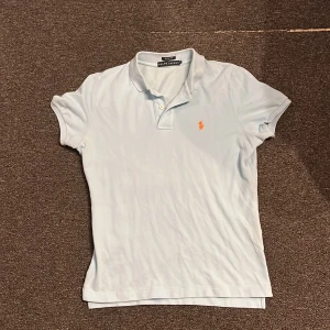 Ljusblå pikétröja från Ralph Lauren - Säljer en stilren ljusblå pikétröja från Ralph Lauren med klassisk krage och knappar. Tröjan har en broderad orange logga på bröstet. Perfekt för en avslappnad men ändå snygg look. Skick 10/10. Skriv för fler frågor 