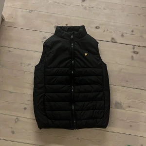 Svart dunväst med dragkedja - Snygg svart dunväst från Lyle & Scott med dragkedja och ett litet broderat emblem på bröstet. Perfekt för lager-på-lager under kyliga dagar.