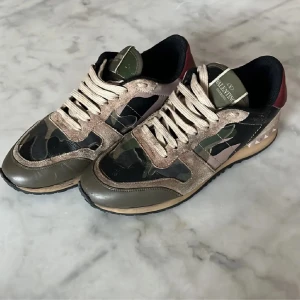 Valentino Rockrunner - Snygga Valentino Rockrunner sneakers med ett coolt camouflage-mönster. Skorna har snörning och är tillverkade i en mix av skinn och mocka. De har en unik design med röda detaljer på hälen och en slitstark sula.