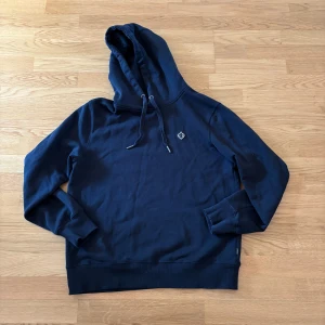Mörkblå hoodie från Morris - Säljer en stilren mörkblå hoodie från Morris. Tröjan har en klassisk design med justerbar huva och ribbade muddar. Perfekt till sommaren och våren
