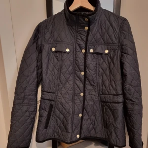 Svart quiltad jacka från Stockh LM - Snygg svart quiltad jacka från Stockh LM med guldknappar och dragkedjor. Jackan har en hög krage och praktiska fickor framtill. Perfekt för kyligare dagar med sin stilrena design. 