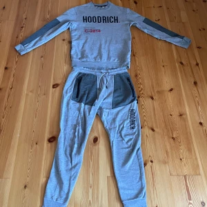 Grå tracksuit från Hoodrich - Säljer ett grå tracksuit från Hoodrich med tryck på både tröja och byxor. Tröjan har långa ärmar och textdetaljer i svart och rött. Byxorna har dragkedjefickor i mörkare grå nyans och elastisk midja med snörning. Perfekt för en avslappnad stil. Både byxorna och tröjan har väldigt snygg baksida. Byxorna har även hoodrich tryck på handtaget på fickorna 