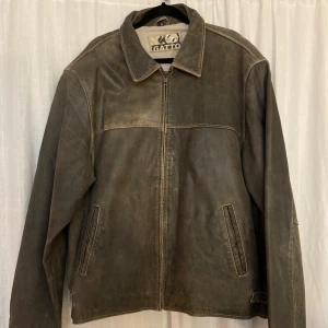 Brun/grå skinnjacka från Gatto - Säljer en brun skinnjacka från Gatto med dragkedja framtill. Jackan har en klassisk krage och två fickor på sidorna. Perfekt för en cool och avslappnad stil. Längd: 71cm armhåla till armhåla: 62cm Axel till Axel: 53cm