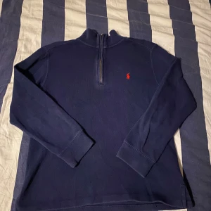 Mörkblå tröja från Ralph Lauren - Säljer en stilren mörkblå tröja från Ralph Lauren i 100% bomull. Tröjan har en dragkedja vid halsen och det klassiska röda broderade logotypen på bröstet. Perfekt för en avslappnad och snygg look.