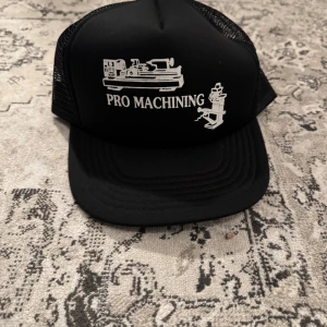 Svart keps med tryck - Snygg svart keps med vitt tryck 'PRO MACHINING' och motiv av maskiner. Kepsen har ett nätmaterial baktill för ventilation och justerbar passform. Kepsen är köpt inne på beyond retro. Aldrig använd.