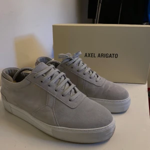 Grå sneakers från Axel Arigato - Snygga grå sneakers från Axel Arigato i mocka med klassisk snörning. Perfekta för en stilren look. Skorna har en vit sula som ger en fräsch kontrast till det gråa.