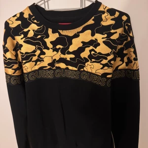 Svart och guld camouflagemönstrad tröja från Guess - Snygg svart tröja från Guess med ett iögonfallande gult camouflagemönster över bröstet. Tröjan har långa ärmar och Guess-loggan tryckt längs med mönstret. Perfekt för en trendig look!