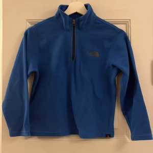 Barn: Blå fleece tröja från The North Face - Säljer en blå fleece tröja från The North Face med halv dragkedja och broderad logga på bröstet. Perfekt för kyliga dagar och utomhusaktiviteter. Tröjan har långa ärmar och en hög krage för extra värme.