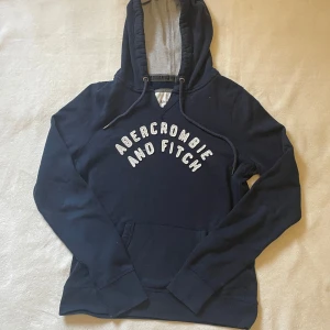 Mörkblå hoodie från Abercrombie & Fitch - Säljer en snygg mörkblå hoodie från Abercrombie & Fitch med vit text på framsidan. Tröjan har en klassisk känguruficka och en bekväm huva med dragsko. Perfekt för en avslappnad stil.