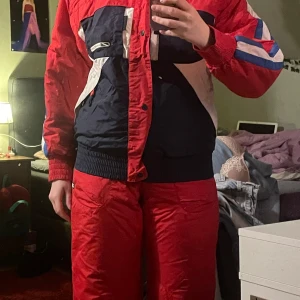 Retro skidjacka och byxor från Ca Cao - Säljer en cool retro skidjacka och matchande byxor från Ca Cao. Jackan är röd, blå och vit med dragkedja och knappar. Den har långa ärmar och flera praktiska fickor. Byxorna är röda med hängslen och dragkedjor vid fickorna. Perfekt för vinterns äventyr i backen! 🎿