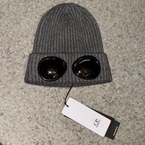 C.P. Company mössa - Snygg grå ribbad mössa från C.P. Company med unika glasögondetaljer framtill. Perfekt för att hålla stilen under kalla dagar. Nyskick och oanvänd. Billig vid snabb affär.