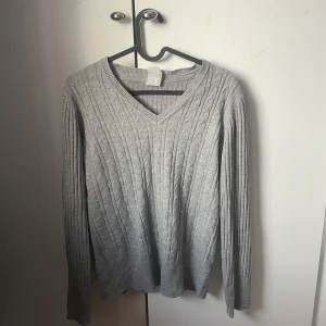 Grå stickad tröja med v-ringning - Säljer en stilren grå stickad tröja med v-ringning. Tröjan har ett klassiskt kabelstickat mönster och långa ärmar. Snygg att styla med ett vitt linne! 