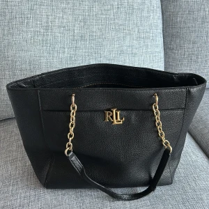 Svart väska från Ralph lauren - Snygg svart axelväska från Ralph Lauren med guldfärgade kedjedetaljer och logotyp. Väskan har en rymlig insida med dragkedja för säker förvaring. Perfekt för att bära med sig alla nödvändigheter med stil.