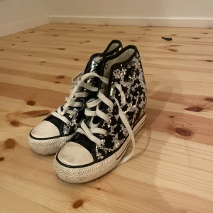 Svarta sneakers med paljetter - Snygga svarta sneakers med vita snören och glittriga paljetter. Perfekta för att ge din outfit en extra touch. De har en klassisk design med högre skaft och vit sula. Skorna har klack
