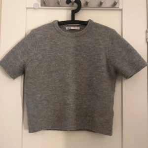 Grå stickad tröja från Zara - Säljer en stilren grå stickad tröja från Zara. Den har korta ärmar och en rund halsringning. Perfekt för lager-på-lager eller som den är.