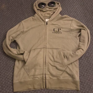 Olivgrön hoodie från C.P. Company - Säljer en olivgrön hoodie från C.P. Company med dragkedja och unika glasögon i huvan. Perfekt för en stilren och avslappnad look. Hoodien har långa ärmar och praktiska fickor framtill. Kan sänka priset vid snabb affär.Är i dem bilderna som visas inte ny tvättad men kommer ny tvättad.
