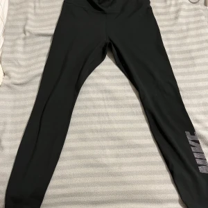 Svarta leggings från Nike - Snygga svarta leggings från Nike med Dri-FIT-teknologi för optimal komfort. De har en hög midja och ett stort Nike-logotyptryck längs benet. Perfekta för träning eller en aktiv vardag. Storlek S