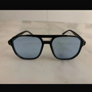 Svarta solglasögon med blå linser - Snygga svarta solglasögon med en modern design och blå tonade linser. De har en dubbel brygga och en klassisk form som passar de flesta ansikten. Perfekta för en stilren look.