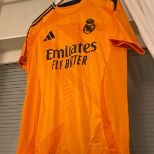 Real Madrid fotbollströja från Adidas - Snygg orange fotbollströja från Adidas med Real Madrid-logga och 'Emirates Fly Better' tryckt på framsidan. Tröjan har korta ärmar och svarta detaljer på axlarna. Perfekt för fotbollsfans som vill sticka ut på planen!