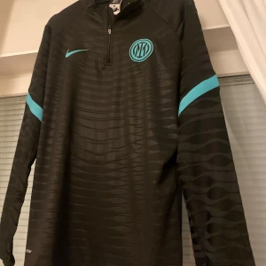 Inter Milan långärmad tröja från Nike - Snygg svart långärmad tröja från Nike med turkosa detaljer och dragkedja vid halsen. Perfekt för fotbollsträningen eller som en stilren vardagströja. Tröjan har ett diskret mönster och klubbemblem på bröstet.