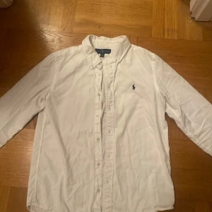 Vit skjorta från Ralph Lauren - Klassisk vit skjorta från Ralph Lauren med broderad logotyp på bröstet. Skjortan har långa ärmar och knappar framtill. Passar dig perfekt som är runt 170
