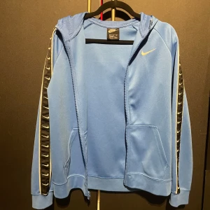 Blå hoodie från Nike - Säljer en blå hoodie från Nike med dragkedja och huva. Den har svarta detaljer längs ärmarna med vita Nike-loggor. Perfekt för en sportig look eller avslappnade dagar. Storlek 170 men passar S