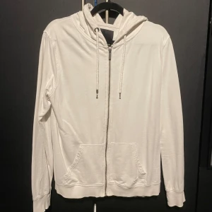 Vit hoodie från 157 - Säljer en stilren vit hoodie från 157 i storlek L men liten i storlek så skulle säga M. Den har en dragkedja framtill och justerbar huva med snören.