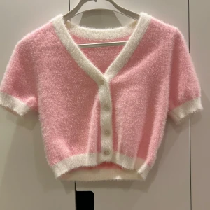 Rosa fluffig kofta med vita detaljer - Säljer en söt rosa kofta med korta ärmar och vita kanter. Den är fluffig och har knappar framtill. Aldrig använd. Vet inte vart den kommer ifrån tyvärr💕