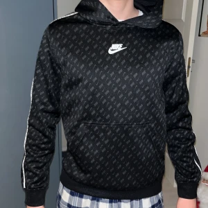 Svart hoodie från Nike - Snygg svart hoodie från Nike med ett diskret mönster och vit logga på bröstet. Tröjan har en huva och vita ränder längs ärmarna. 