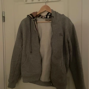 Grå hoodie från Burberry - Snygg grå hoodie från Burberry med dragkedja och klassiskt rutigt mönster i huvan. Tröjan har långa ärmar och praktiska fickor framtill. Perfekt för en stilren och bekväm look. Använd men inga tydliga tecken. Storlek S