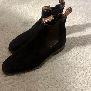 Svarta Chelsea boots från R.M. Williams - Snygga bruna Chelsea boots från R.M. Williams i mocka. De har en klassisk design med elastiska paneler på sidorna och dragflikar för enkel påtagning. Perfekta för en stilren look.