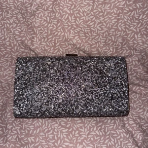 Glittrig kuvertväska - Snygg glittrig kuvertväska i silver med en elegant design. Perfekt att ha på balen eller bara för att lägga till lite bling till din outfit. Väskan har en metallram och en praktisk stängning upptill. Använd en gång.