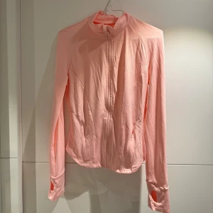 Rosa träningsjacka med dragkedja - Säljer en ljusrosa jacka med dragkedja och långa ärmar. Jackan har praktiska tumhål vid ärmsluten och två fickor med dragkedja framtill. Perfekt för en sportig look eller en avslappnad dag.