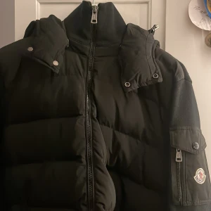 Äkta moncler cardigan - Snygg svart dunjacka från Moncler med avtagbar huva och dragkedja framtill. Jackan har en praktisk ficka med dragkedja på ärmen och tryckknappar vid kragen. Perfekt för kyliga dagar.