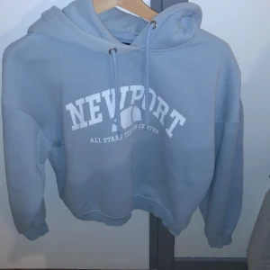 Blå hoodie från 157 - Säljer en blå hoodie från 157 med texten 'Newport All Stars Tennis Center' tryckt i vitt på framsidan. Hoodien har en klassisk design med dragsko i huvan och ribbade muddar vid ärmslut och nederkant.