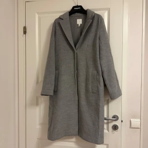 Grå kappa från H&M - Snygg ljudgrå kappa från H&M i en klassisk design. Den har långa ärmar och två praktiska fickor framtill. Perfekt för kyligare dagar och ger en stilren look.