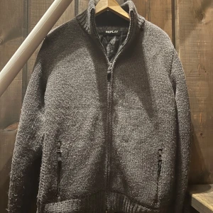 Grå stickad jacka från Replay - Snygg grå stickad jacka från Replay med dragkedja och ribbade detaljer. Perfekt för kyliga dagar med sin mysiga och stilrena design. Jackan har praktiska fickor med dragkedja och en hög krage för extra värme.