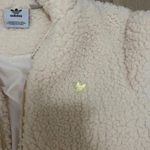 Beige teddyjacka från Adidas - Mysig beige teddyjacka från Adidas med huva och dragkedja. Tillverkad i 100% . Jackan har en broderad Adidas-logga på bröstet och ribbade muddar vid ärmslut och nederkant.
