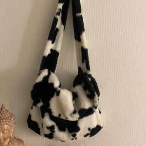 Svartvit axelväska med pärldetalj - Snygg och unik axelväska i svartvitt mönster som påminner om ko-mönster. Väskan har en mjuk och fluffig textur med en fin pärldetalj på ”öronen”. Perfekt för att ge din outfit en cool och trendig touch.