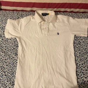Vit pikétröja från Ralph Lauren - Säljer en klassisk vit pikétröja från Ralph Lauren med broderad logga i blått på bröstet. Tröjan är kortärmad och passar inte min stil så varför inte sälja den. Har Max använt den 1-2 gånger.