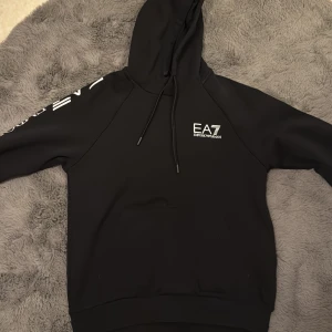 Svart hoodie från Emporio Armani - Snygg svart hoodie från Emporio Armani med EA7-logga på bröstet och ärmen. Den har en klassisk design med dragsko i huvan och en stor ficka framtill. Perfekt för en stilren look.