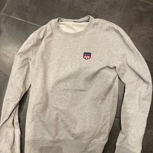 Grå sweatshirt från GANT - Säljer en stilren grå sweatshirt från GANT med ett broderat emblem på bröstet. Tröjan har långa ärmar och en rund halsringning. Perfekt för en avslappnad look.