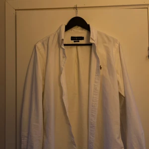 Vit skjorta från Ralph Lauren - Säljer en klassisk vit skjorta från Ralph Lauren i slim fit. Skjortan har långa ärmar och är tillverkad i bomull. Den är dekorerad med det ikoniska broderade logotypen på bröstet. Perfekt för en stilren look.