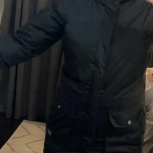 Svart dunjacka från Everest - Säljer en svart dunjacka från Everest med dragkedja och knappar. Jackan har långa ärmar och praktiska fickor framtill. Perfekt för kalla dagar.