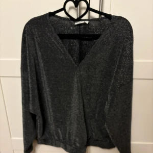 Glittrig svart blus - Snygg glittrig svart blus med v-ringning och långa ärmar. Perfekt för en festlig kväll eller en utekväll med vänner. Blusen har en lös passform som ger en bekväm känsla.